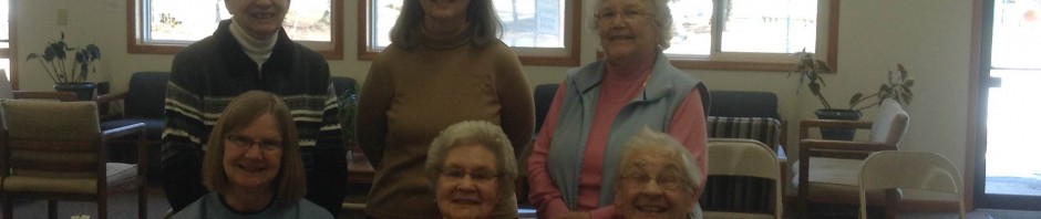 Group of six women smiling with handmade quilts in a cozy room.