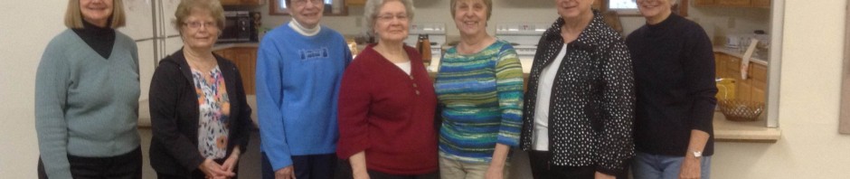 Seven women standing in a row inside a room with shelves and a sign.