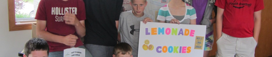 Group of boys holding a lemonade stand sign indoors.