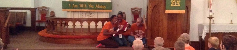 Children seated on a church floor near the altar, adults seated in pews.