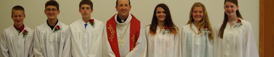 A group of young people and a priest in white robes, standing indoors.