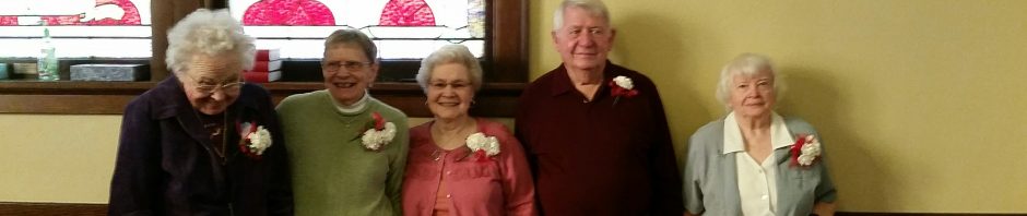 Five elderly people standing indoors near a stained glass window.