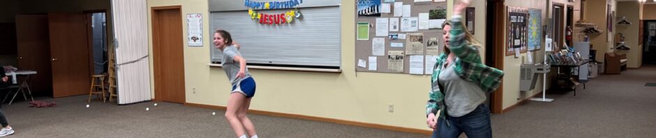 Two people in a gym practicing tennis with rackets and a ball.