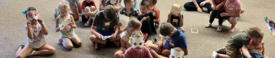 Children wearing masks sit on the floor in a classroom setting during an educational event.