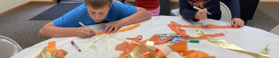 Three children crafting with colorful paper strips at a table indoors.