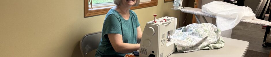 Woman sewing at a table with a sewing machine near a window.
