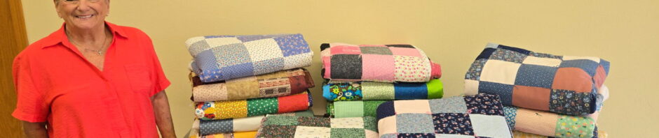 Stacks of colorful patchwork quilts on a table against a yellow wall.